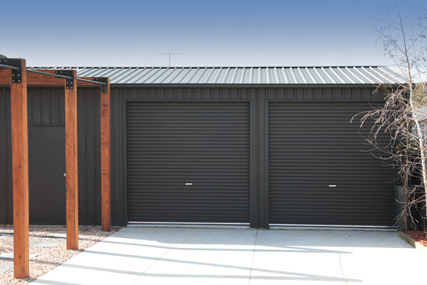 Black shed in COLORBOND steel Monument Matt with two roller doors, personal access door to left and wooden pergola in front
