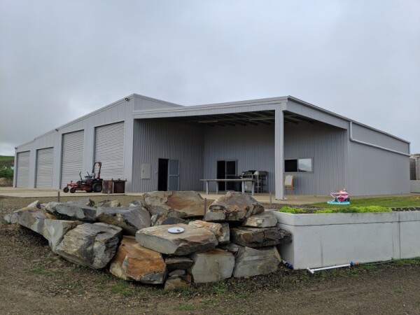 Tailor-made steel shed with four garage doors to the left, an open bay on a lean-to to the right and a retaining wall and rock feature in the foreground