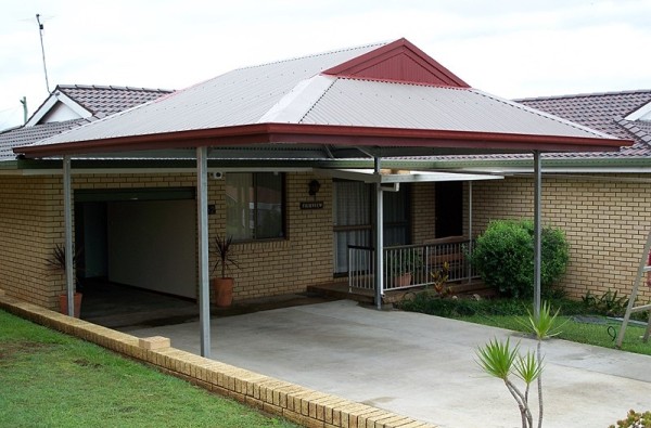 A residential carport with a dutch gable steel roof over a concrete driveway on a brick house